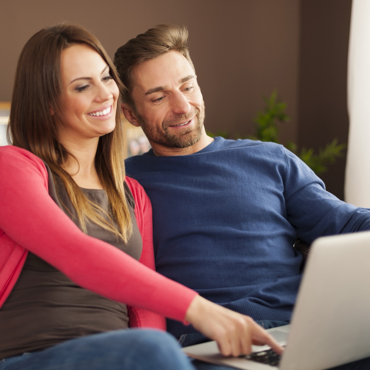 Smiling couple on the sofa at home, happily browsing property listings on a laptop